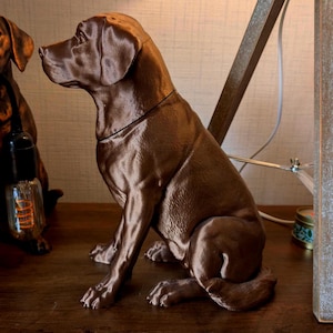 May include: A brown dog statue in a seated position, with a smooth, glossy finish. The dog has detailed features, including its ears, paws, and tail. The statue is on a wooden surface, with a lamp and another dog statue in the background.