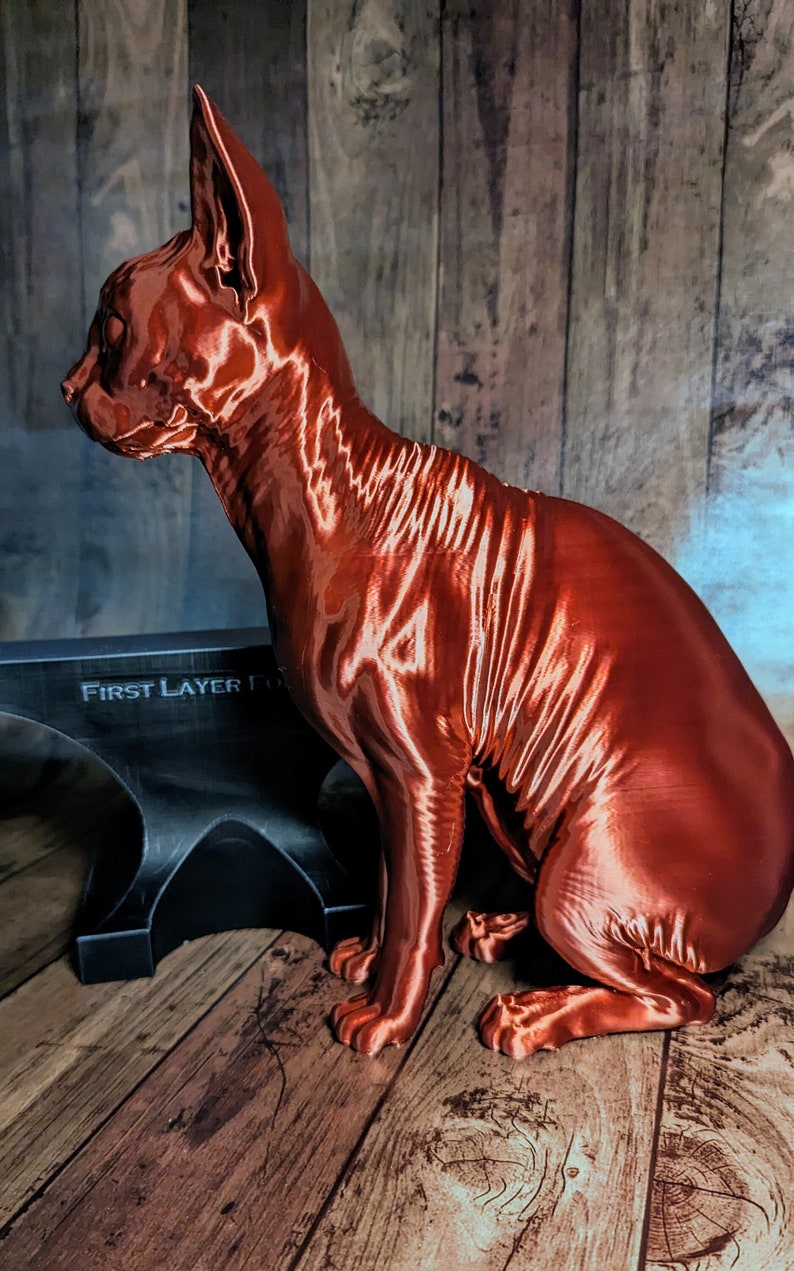 Op de afbeelding: Een 3D-geprint koperkleurig kattenbeeldje dat op een houten oppervlak staat. De kat is haarloos en heeft een gladde, glanzende afwerking.