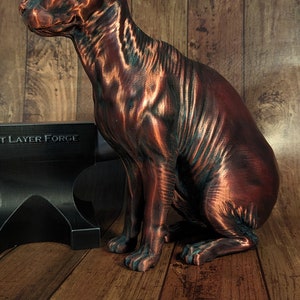 Op de afbeelding: Een koperkleurig, 3D-geprint beeld van een haarloze kat die op een houten oppervlak zit. Het beeld is gedetailleerd en realistisch, met zichtbare rimpels en plooien in de huid van de kat. Het beeld staat op een houten oppervlak met een zwarte metalen aambeeld op de achtergrond. Het aambeeld heeft de tekst "3D Layer Forge" erop gedrukt.