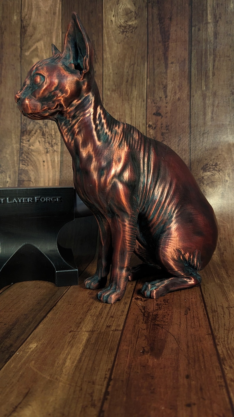 Op de afbeelding: Een koperkleurig, 3D-geprint beeld van een haarloze kat die op een houten oppervlak zit. Het beeld is gedetailleerd en realistisch, met zichtbare rimpels en plooien in de huid van de kat. Het beeld staat op een houten oppervlak met een zwarte metalen aambeeld op de achtergrond. Het aambeeld heeft de tekst "3D Layer Forge" erop gedrukt.