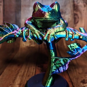 May include: A metallic frog sculpture perched on a branch with leaves. The frog and branch have a color-shifting finish, displaying hues of green, blue, and purple. The sculpture is set against a wooden background.