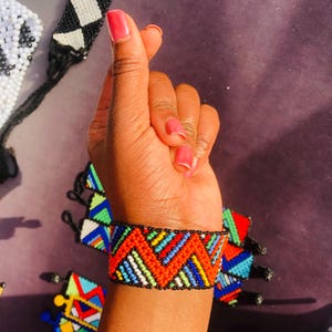 May include: A wide cuff bracelet crafted from small glass beads in a vibrant geometric pattern. The design features repeating zigzag lines in bright red, blue, green, yellow, and white, bordered by rows of tiny black beads along the top and bottom edges.