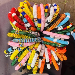 May include: A collection of circular bracelets made from small, multi-colored glass beads. The bracelets feature various patterns and color combinations, including red, yellow, blue, green, orange, pink, black, and white, arranged in a dense cluster.