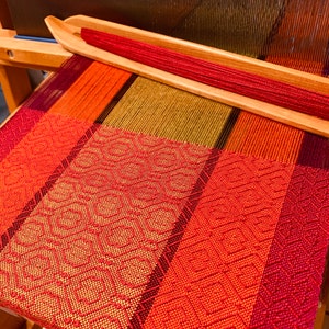 May include: A loom with a woven fabric in shades of red, orange, and green. The fabric is being woven on a wooden loom with a red yarn shuttle.