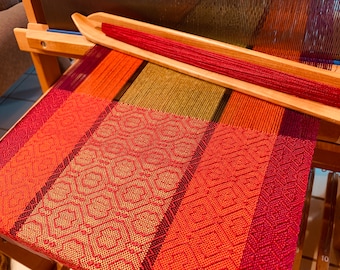 May include: A loom with a woven fabric in shades of red, orange, and green. The fabric is being woven on a wooden loom with a red yarn shuttle.