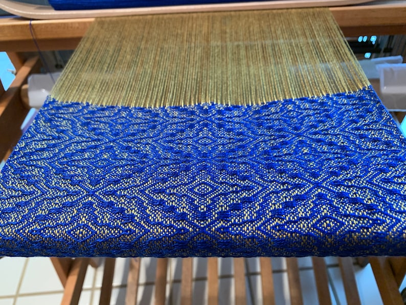 May include: A close-up of a loom with a blue and gold woven fabric. The fabric has a geometric pattern.