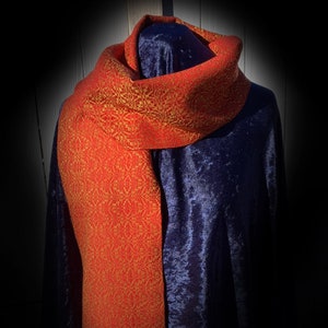 May include: A red and gold woven scarf with a geometric pattern. The scarf is draped over a dark blue velvet surface.