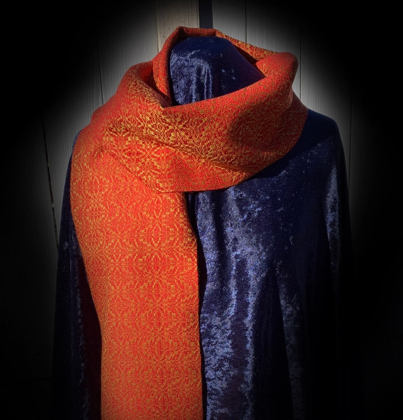 May include: A red and gold woven scarf with a geometric pattern. The scarf is draped over a dark blue velvet surface.