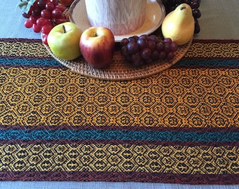 May include: A patterned table runner in gold, teal, and burgundy. The runner has a geometric design with repeating diamond and floral motifs. Fresh fruit, including apples, grapes, and a pear, sits on a woven plate. The runner is rectangular.