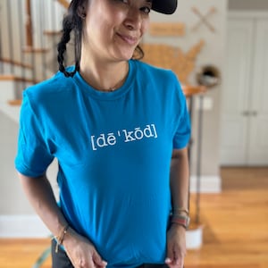 Peut inclure: Une femme portant un t-shirt bleu turquoise avec le texte "[de'kod]" en lettres blanches. Elle porte également une casquette de baseball noire et rouge et une jupe noire.