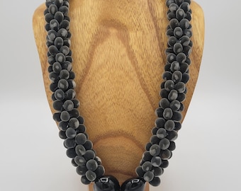 May include: A dark gray necklace with a braided design, displayed on a wooden jewelry bust. The necklace features small, rounded beads and two larger, glossy black beads at the bottom. The background is a plain white.