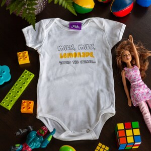 May include: White baby onesie with the text "milk, milk, lemonade, 'round the corner..." in yellow lettering. The onesie is surrounded by toys, including a doll, blocks, and a Rubik's cube.