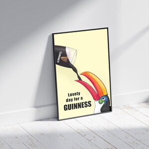 K&ouml;nnte beinhalten: Ein schwarzer gerahmter Poster mit gelbem Hintergrund. Ein Tukan mit rotem und orangefarbenem Schnabel trinkt aus einem Glas Guinness-Bier. Der Text auf dem Poster lautet "Lovely day for a GUINNESS".