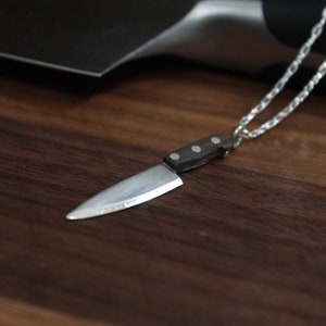 May include: A silver chain necklace with a miniature knife pendant. The knife has a silver blade and a brown wooden handle.