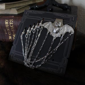 May include: A silver-toned bat-shaped bookmark with dangling chains. The bookmark is attached to a dark, patterned surface, with the wings of the bat spread wide. The bookmark is a gothic-themed accessory.