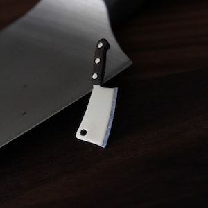 May include: A miniature cleaver with a white blade and a dark wooden handle with three white rivets. The cleaver is set against a larger, blurred metal blade, all on a dark wood surface.