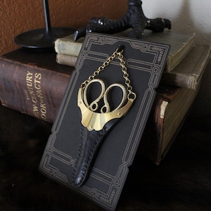 May include: A gold-colored pair of scissors with a black leather sheath and a chain. The scissors are displayed on a black card with a decorative border.