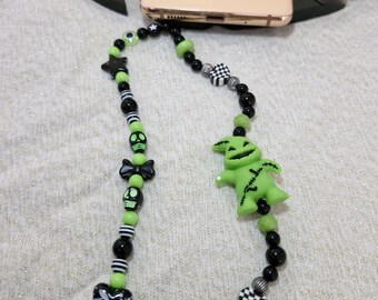 May include: A beaded phone strap featuring black, green, and white beads with skull, star, and heart charms. The strap includes a green Oogie Boogie charm. A gold phone with the text "Poison Peach" rests above the strap.