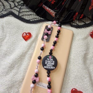 May include: A phone strap with black, pink, and clear beads, and a circular charm with the text "I'M TOO CLUMSY TO BE AROUND FRAGILE MASCULINITY". The strap is draped over a phone. Red heart-shaped embellishments are scattered around.