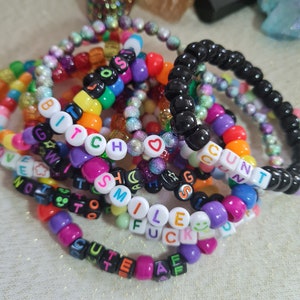 May include: A close-up of several colourful beaded bracelets with words spelled out on them, including "BITCH", "SMILE", "FUCK", "CUTE", and "GUNT".