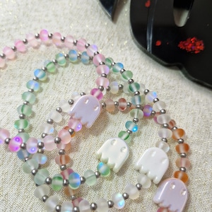 May include: Three beaded bracelets with iridescent glass beads in pastel colours, featuring small ghost-shaped charms. The bracelets have silver-coloured metal accents. A black decorative object is in the background.
