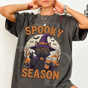 Puede incluir: Una camiseta gris carbón con el gráfico de un gato negro con sombrero de bruja morado y gafas, sosteniendo una taza de café. El gato está frente a una luna llena y siluetas de árboles. El texto "SPOOKY SEASON" está impreso en letras naranjas.