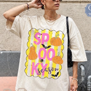 Puede incluir: Una camiseta color hueso con un gráfico de rayas verticales amarillas y un borde festoneado. El diseño incluye calabazas naranjas, una araña negra, una telaraña y dos murciélagos negros. El texto dice "SPOOKY season" en rosa y negro.