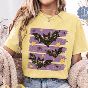 Puede incluir: Una camiseta de manga corta de color amarillo pálido con un estampado gráfico de tres murciélagos negros con grandes ojos amarillos, pequeñas arañas negras y estrellas amarillas sobre rayas horizontales moradas en el frente.