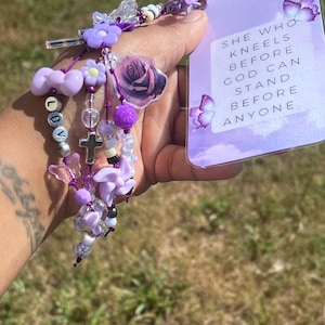 May include: A purple beaded charm bracelet with flower and heart accents, a cross, and the word "LOVE" spelled out in beads. A card with the text "SHE WHO KNEELS BEFORE GOD CAN STAND BEFORE ANYONE" is also visible.
