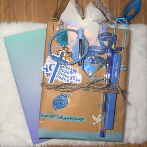 Könnte beinhalten: Ein Geschenkset, verpackt in braunem Papier, mit Bindfaden verschnürt und mit blauen Akzenten verziert. Enthält ein Notizbuch mit blau-violettem Farbverlauf, Stifte, ein Armband und Aufkleber mit religiösen Themen. Das Set ist mit einem blauen Band und einer weißen Schleife verziert.