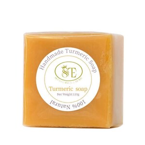 May include: A square bar of yellow turmeric soap with a white label that reads "Handmade Turmeric Soap" and "100% Natural" with a logo of a crown and the letters "SE" inside a circle. The label also says "Turmeric Soap" and "Net Weight: 110g".