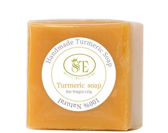 May include: A square bar of yellow turmeric soap with a white label that reads "Handmade Turmeric Soap" and "100% Natural" with a logo of a crown and the letters "SE" inside a circle. The label also says "Turmeric Soap" and "Net Weight: 110g".