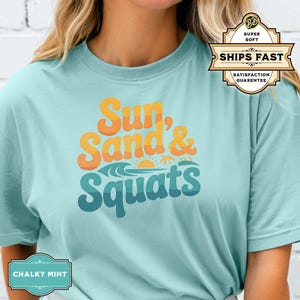 May include: A light blue t-shirt with the text "Sun, Sand & Squats" in a retro font. The text is in shades of yellow, orange, and blue. The shirt has a relaxed fit and short sleeves.