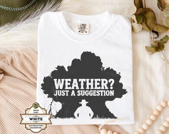 Op de afbeelding: Wit T-shirt met een zwarte opdruk met de tekst "WEATHER? JUST A SUGGESTION". Het ontwerp toont de silhouet van een persoon met een cowboyhoed. Het shirt heeft een ronde hals en is gemaakt van een zacht materiaal.