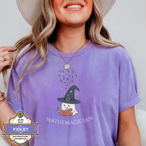 May include: A purple t-shirt with a graphic of a ghost wearing a wizard hat and holding a book. Above the ghost are math symbols. The word "MATHEMAGICIANS" is printed below the ghost.