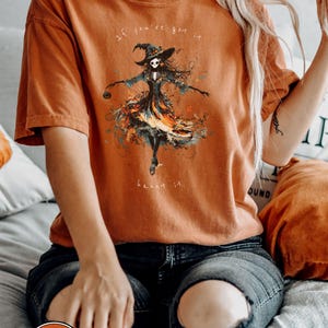 A spooky yet chic t-shirt design featuring a dancing skeleton girl in a flowing gown, holding a jack-o-lantern, with the phrase If You've Got It, Haunt It. Perfect for Halloween enthusiasts, gothic fashion fans, and stylish Halloween events.