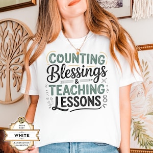 May include: White t-shirt with the text "COUNTING BLESSINGS & TEACHING LESSONS" in black, green, and light yellow. The shirt has a crew neck and short sleeves. The design includes a pencil and other small graphics.