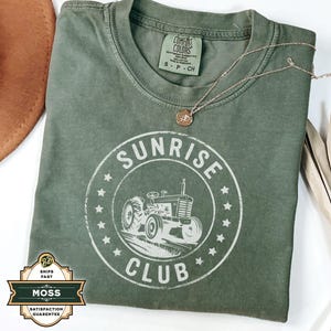 May include: An olive green t-shirt featuring a circular graphic with a tractor illustration and the words "SUNRISE CLUB". A gold necklace with a pendant lies on the shirt. The shirt has a Comfort Colors tag. A brown hat and dried plant are in the background.