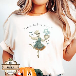 May include: Ivory-colored t-shirt with a whimsical Halloween-themed graphic. The design features a skeleton chef holding a coffee cup with a ghost, surrounded by bats and coffee beans. The text "Death Before Decaf" is above the graphic.