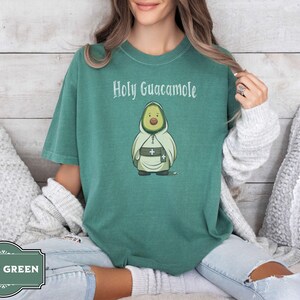 A humorous t-shirt design featuring an avocado dressed as a monk with a cross belt, perfect for food humor enthusiasts. Ideal for those who love avocado puns, funny Christian gifts, and funny phrase slogans for a unique twist on Holy Guacamole.