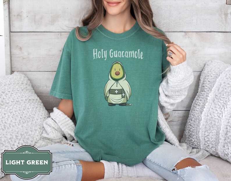 A humorous t-shirt design featuring an avocado dressed as a monk with a cross belt, perfect for food humor enthusiasts. Ideal for those who love avocado puns, funny Christian gifts, and funny phrase slogans for a unique twist on Holy Guacamole.