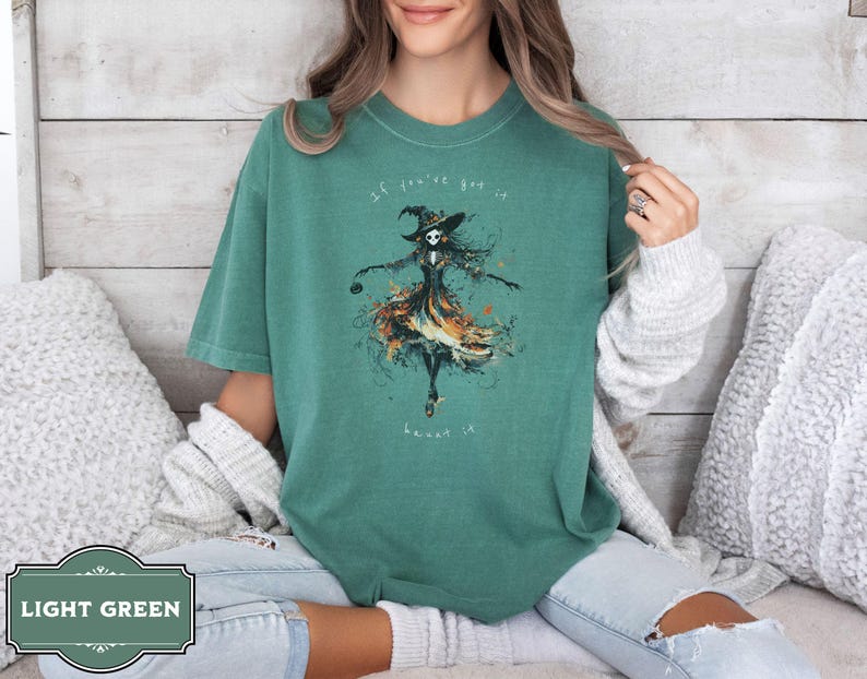 A spooky yet chic t-shirt design featuring a dancing skeleton girl in a flowing gown, holding a jack-o-lantern, with the phrase If You've Got It, Haunt It. Perfect for Halloween enthusiasts, gothic fashion fans, and stylish Halloween events.