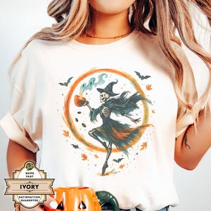 May include: Ivory-colored t-shirt featuring a Halloween-themed graphic. The design includes a skeleton wearing a witch hat and cape, holding a cup with steam rising. The graphic is set within an orange circle with bats and leaves.