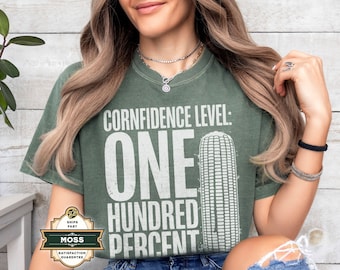 Op de afbeelding: Groen t-shirt met de tekst "CONFIDENCE LEVEL: ONE HUNDRED PERCENT" en een afbeelding van maïs. Het shirt heeft een gedempte groene kleur en een vintage look. De tekst is wit.