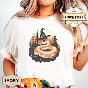 Peut inclure: Un t-shirt blanc avec un graphique représentant un serpent portant un chapeau de sorcière dans un décor de forêt. Le serpent est orange et brun avec un chapeau noir. Le texte "IVORY" est visible dans le coin inférieur gauche de l'image.