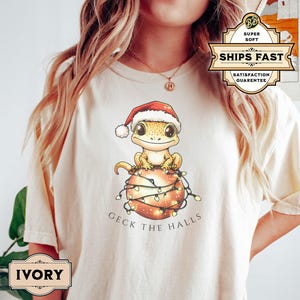 May include: A white t-shirt with a cartoon gecko wearing a Santa hat and sitting on a Christmas ornament with lights. The text "Geck the Halls" is printed below the image.