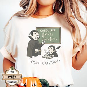 May include: Ivory-colored t-shirt featuring a cartoon vampire teaching calculus. The chalkboard displays mathematical equations. Below the image is the text "COUNT CALCULUS". The shirt has rolled-up sleeves.