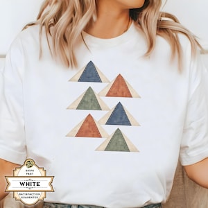 May include: A white short-sleeve t-shirt featuring a graphic print of six stacked triangles in muted shades of blue, green, and terracotta. Each triangle has a darker inner section with a visible stitched border, arranged in a scattered pattern.