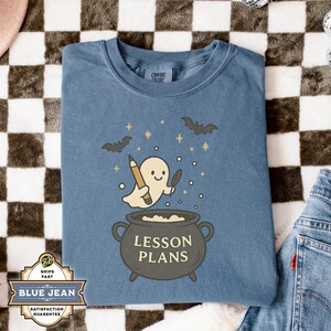 May include: Blue t-shirt with a Halloween-themed graphic. The design features a friendly ghost holding a pencil and a knife above a cauldron that says "LESSON PLANS". Bats and stars are also included in the design.
