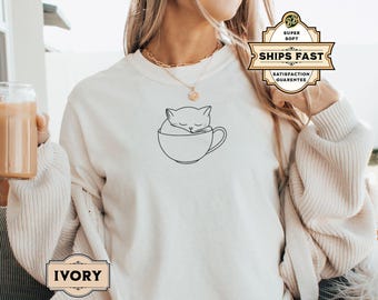 May include: A white sweatshirt with a black line drawing of a cat sleeping in a teacup. The text "IVORY" is on a label in the bottom left corner of the image.